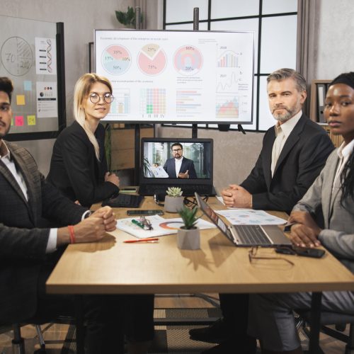 Portrait of group of four skillful multiethnical businesspeople looking at the camera while having video conference from laptop in boardroom, with their male colleague explaining some financial charts